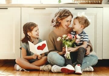 a mum sitting on the ground with two kids who are giving her a mothers day card.